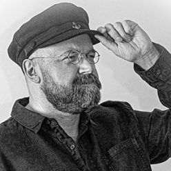 A bearded man in glasses holding the rim of his sailor's cap